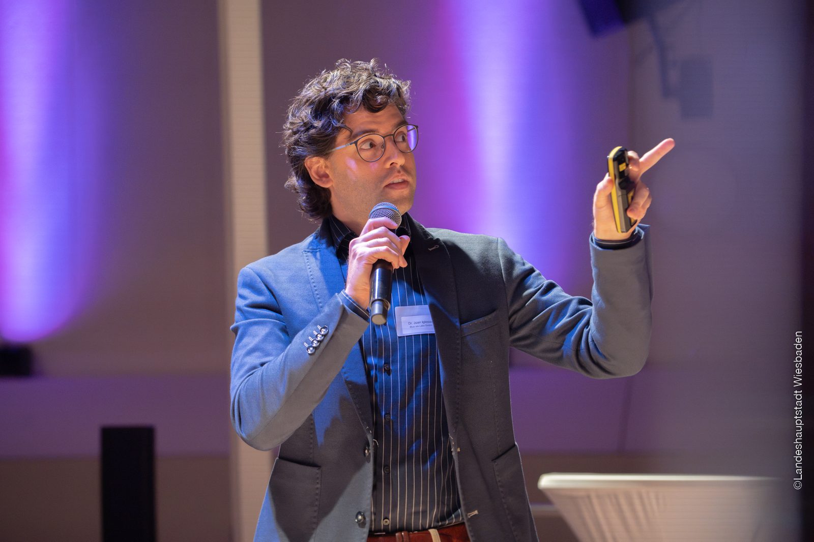 Juan Iglesias Artola on stage with a microphone in one hand, pointing with the other towards the screen behind him, under violet stage lighting.