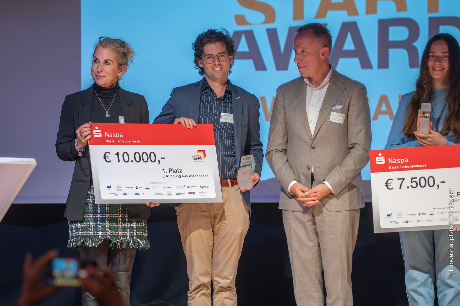 Juan Iglesias Artola on stage holding the first-place trophy and the prize cheque for “Gründung aus Wiesbaden”, between two presenters, in front of the StartAward logo.