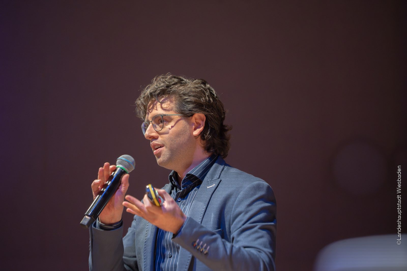 Juan Iglesias Artola in profile against a dark backdrop, speaking into a handheld microphone and holding a presentation clicker.