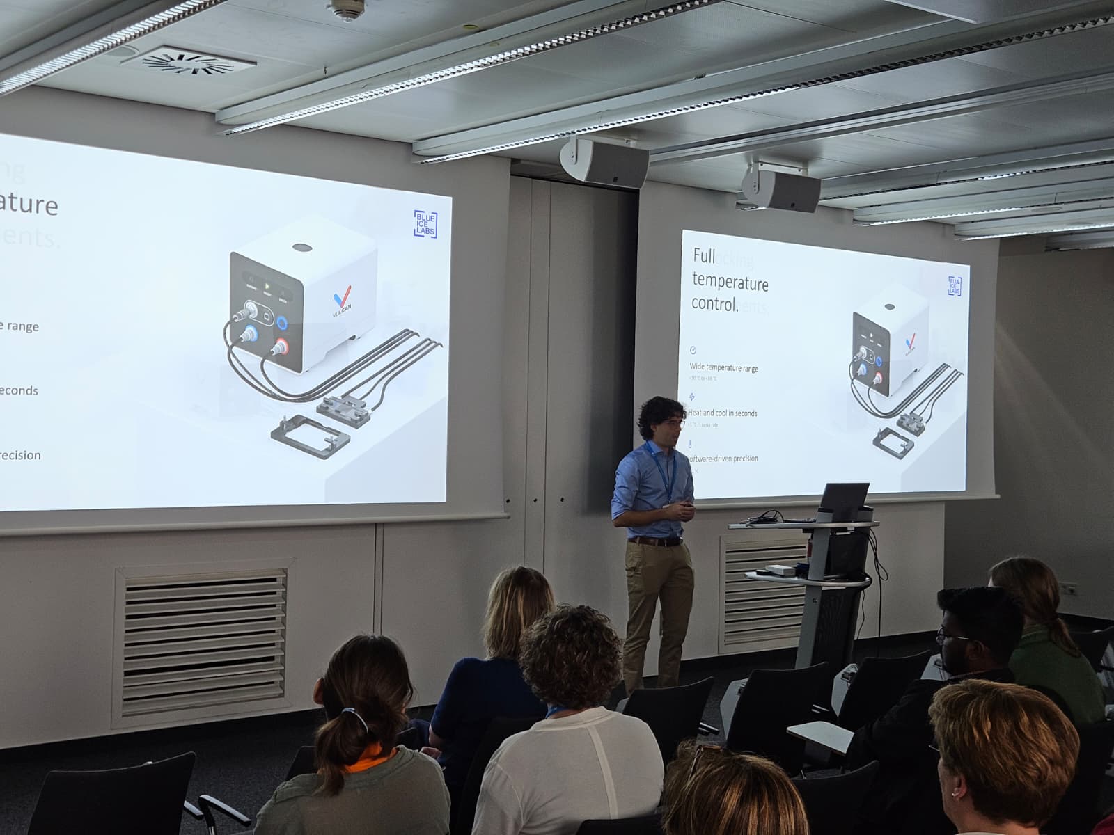 Juan Iglesias presenting to a seated audience. Two screens show a slide headed “Full temperature control”, with a rendering of the control unit and the parts it drives.
